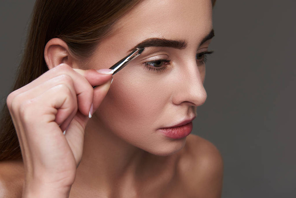 Beautiful young woman carefully plucking eyebrows with tweezers - Фото, изображение