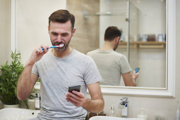 Man brushing his teeth and using mobile phone in bathroom - Fotó, kép