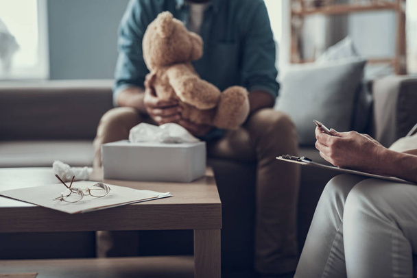 Man sitting on the sofa with teddy bear and his psychotherapist holding notes on the laps - Foto, Imagem