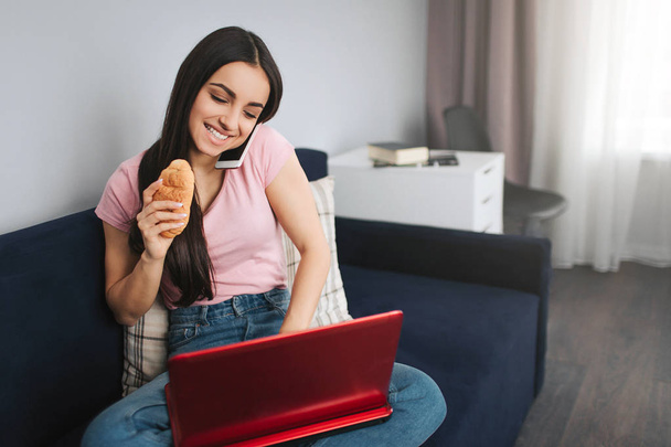 Cheerful young woman work at home. She sit on sofa and type on laptop keyboard. Model talk on phone and hold croissan in hand. - Foto, afbeelding
