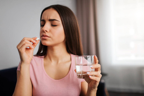 Young woman hold white pill in hand. She is going to eat it. Model hold glass of waterin hand. She looks upset and depressed. - Photo, Image