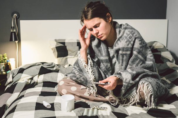 Тематические заболевания и лечение. молодая белая женщина сидит дома спальня кровать завернутая в одеяло тела с высокой температурой, лихорадка держит сильную головную боль за головой, использует капли лекарства в нос от синусита
 - Фото, изображение