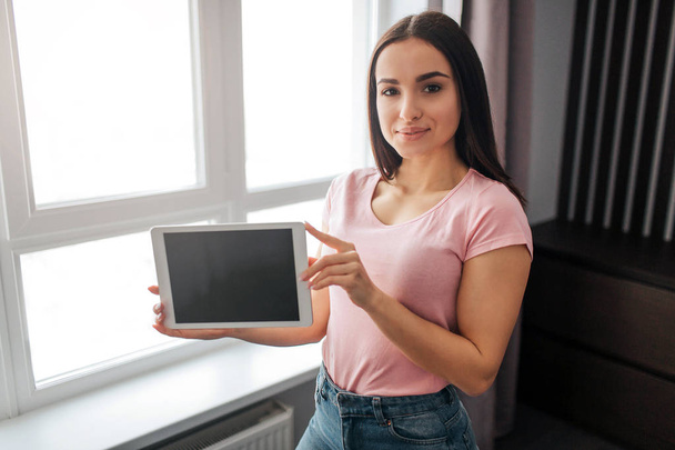 Nice and confident young woman stand at window and look on camera. She hold tablet with dark screen. Model smile a bit. She is in room. - Fotografie, Obrázek