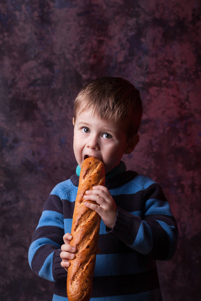 Χαριτωμένο παιδί κρατώντας και δαγκώνει γαλλικό ψωμί ενάντια σε σκούρο κόκκινο φόντο. Αγόρι με όρεξη τρώει ψωμί. - Φωτογραφία, εικόνα