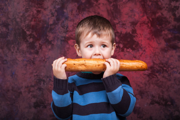 Милий дитина тримає і кусає французький хліб на темному тлі червоною. Хлопчик з апетитом їсть хліб. - Фото, зображення