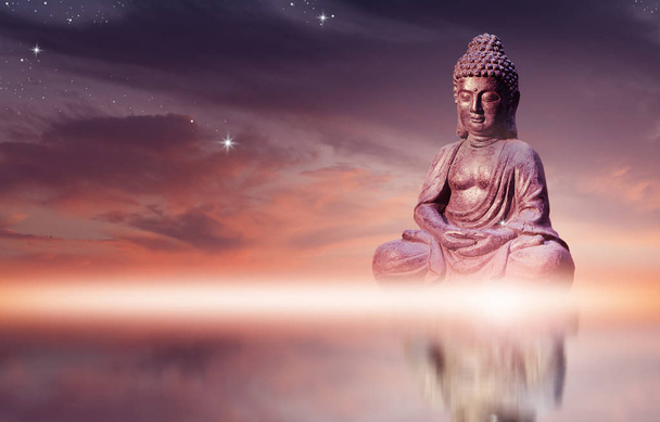 Buddha statue sitting in meditation pose against sunset sky with golden tones clouds. - Photo, Image