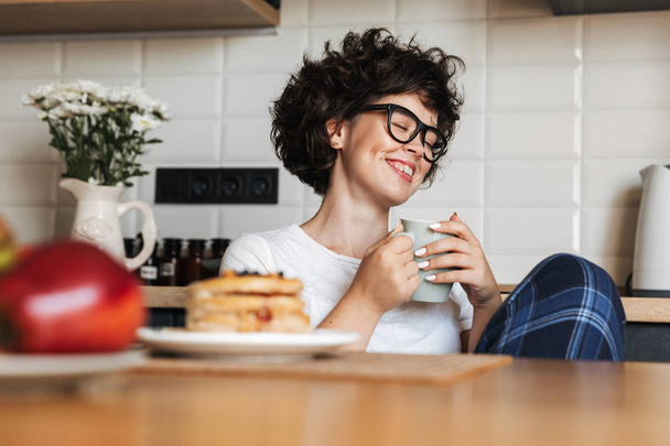 Smiling cheerful girl having tasty breakfast while sitting at the kitchen at home, drinking coffee - Photo, Image