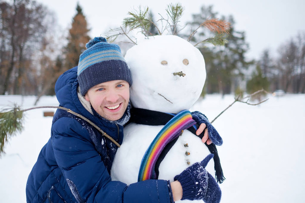 Young european man in warm clothes making snowman from snow outdoor. Having fun like a child at winter vacation. - Фото, изображение