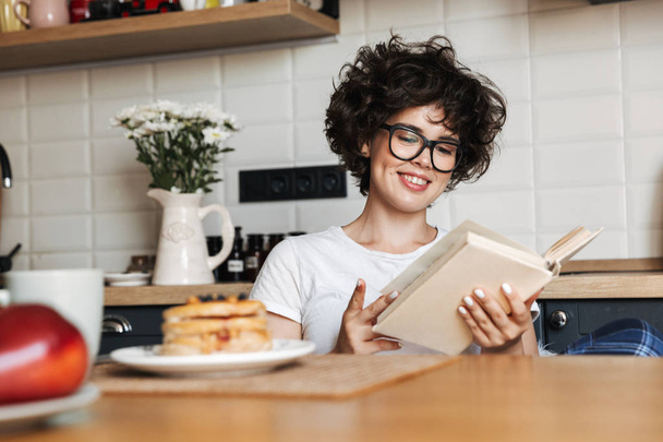 Smiling cheerful girl having tasty breakfast while sitting at the kitchen at home, reading a book - 写真・画像
