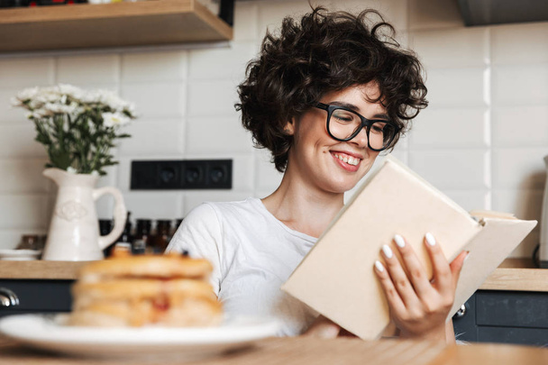 Smiling cheerful girl having tasty breakfast while sitting at the kitchen at home, reading a book - Photo, image