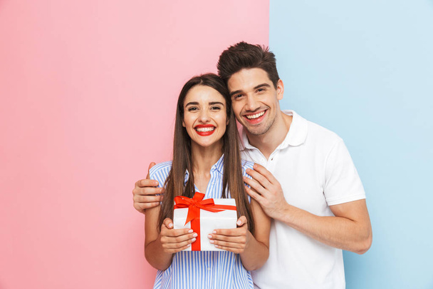 Gelukkige jonge paar staande geïsoleerd over twee gekleurde achtergrond, vieren, houden van de doos van de gift - Foto, afbeelding