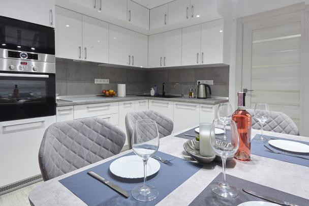 White kitchen interior with grey chairs and served table with white plates glasses and moss on the wall in new luxury home with lights on - Photo, Image