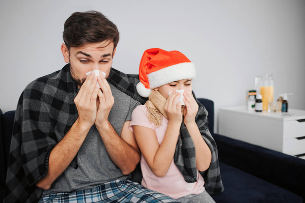 Sick young man and small girl sneezing into white napkin. They caught cold. Girl wear Christmas hat. Family sit in one room on bed. - Photo, Image