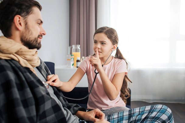 Small girl listen to dads breathing through stethoscope. She hold finger on lips and look at him. Sick young man covered with blanket and scarf. - Fotoğraf, Görsel
