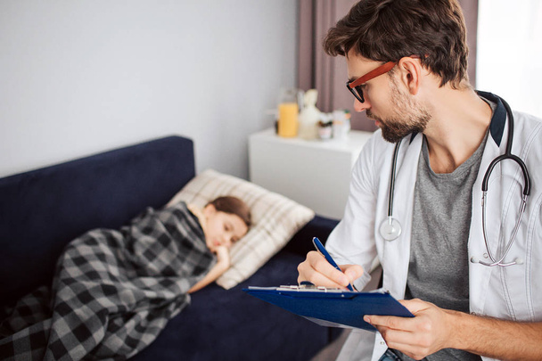 Sick small girl lying on bed covered with blanket. Young male doctor look at her and write. Stethoscope hanging around his neck. - Foto, Bild