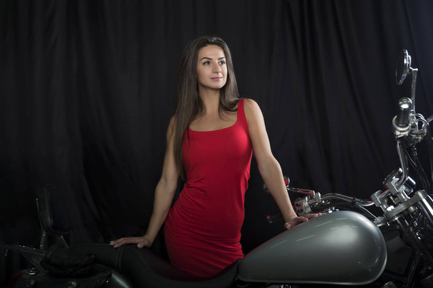 A beautiful young brunette in a bright red dress stands near a chrome motorcycle on a black background - Photo, Image