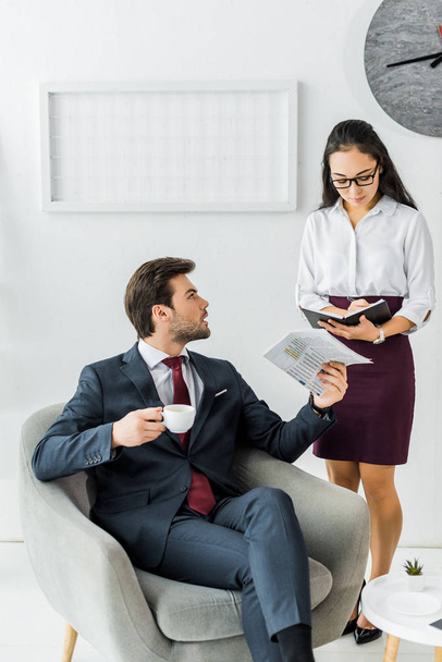 asian businesswoman writing in notebook while businessman sitting on armchair with newspaper and coffee in office - Photo, Image