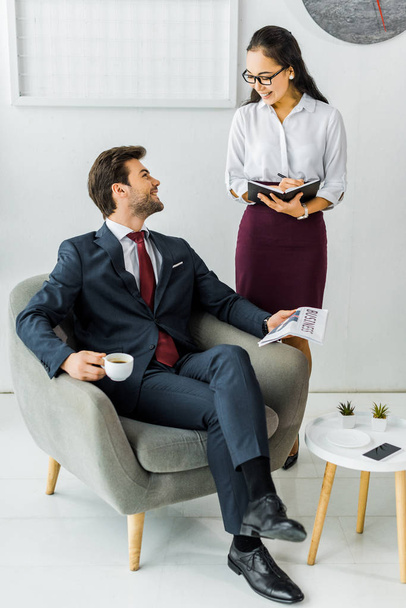 asiatique femme d'affaires écriture dans notebook tandis que l'homme d'affaires assis sur fauteuil avec journal et café au bureau
 - Photo, image