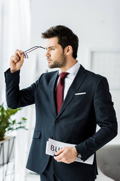 handsome businessman in formal wear holding newspaper and putting on glasses in office - Foto, imagen