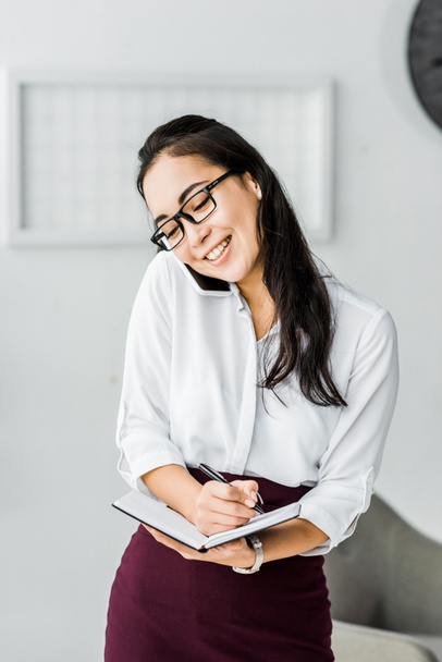 smiling asian businesswoman talking on smartphone and writing in notebook in office - Photo, image