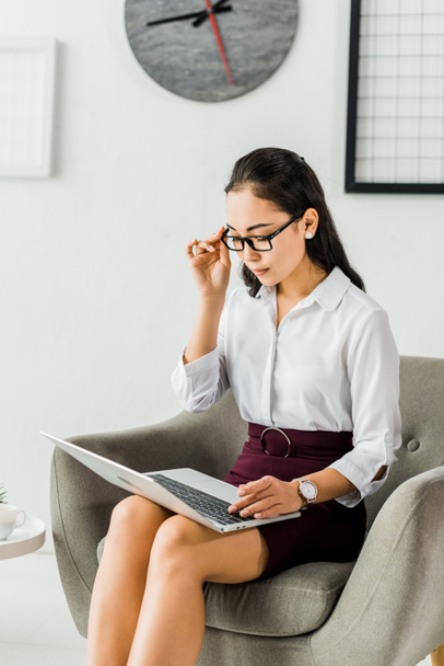 belle asiatique femme d'affaires dans des lunettes assis sur fauteuil et en utilisant ordinateur portable au bureau
 - Photo, image