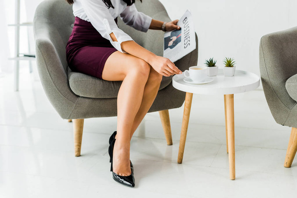 vista recortada de la mujer de negocios sentada en el sillón y la celebración de periódico con taza de café en la oficina
  - Foto, imagen