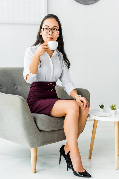 beautiful asian businesswoman in glasses sitting on armchair and having coffee break in office - Фото, изображение