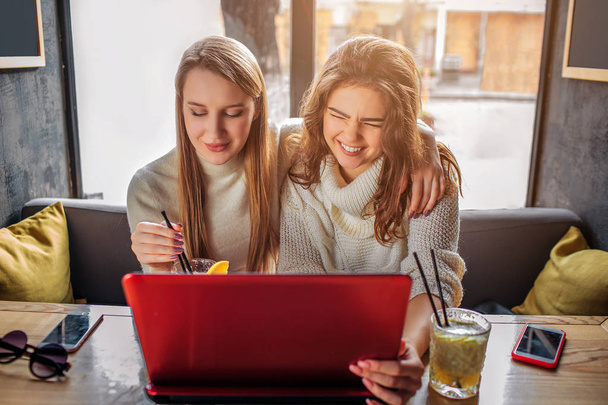 Nice and cheerful young women sit inside at table and watch on laptop. One of them laughing. There are phones and drinks on table. - 写真・画像
