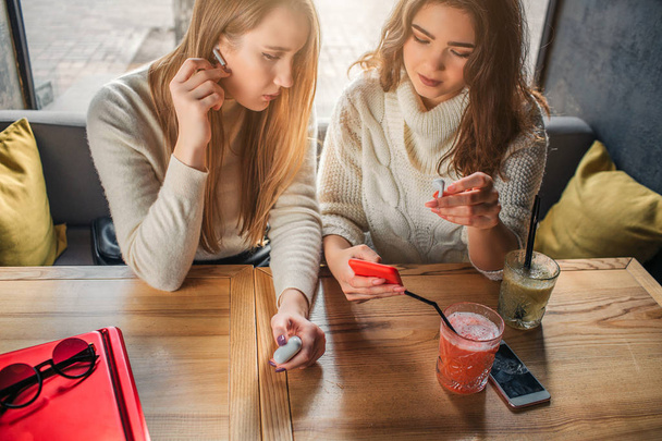 Young women sit at table. Brunette point at phone she has in hands. Blonde hold finger on wireless heaphones. They are concentrated. Model on right talk. - Foto, Bild
