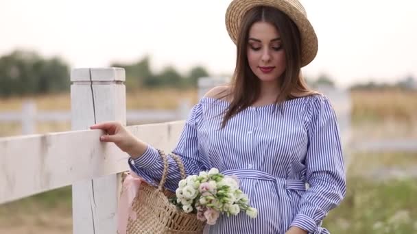 Красивая беременная женщина в синем платье ходит слышать ферму. Вязаная сумка с букетом цветов. Фон дерева и гор. Медленное движение
. - Кадры, видео