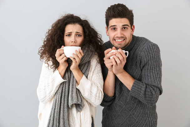 Frozen couple wearing sweaters standing isolated over gray background, holding cups - Foto, immagini