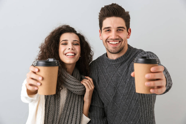 Cheerful couple wearing sweaters standing isolated over gray background, showing takeaway coffee cups - Photo, image