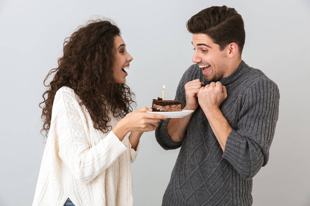 Alegre pareja vistiendo suéteres de pie aislados sobre fondo gris, celebrando cumpleaños, sosteniendo pedazo de pastel con vela
 - Foto, Imagen