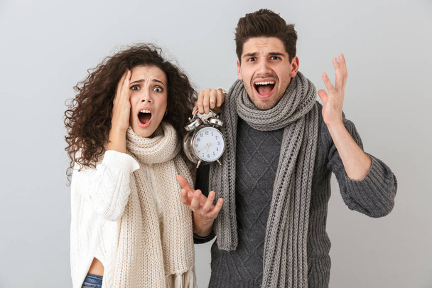 Image of upset couple man and woman screaming while holding alarm clock isolated over gray background - 写真・画像