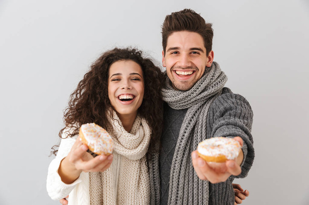 Cheerful young couple wearing sweaters and scarfs standing isolated over gray background, showing donuts - Фото, изображение