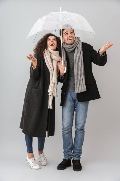 Cheerful young couple wearing sweaters and scarfs isolated over gray background, standing under an umbrella - Photo, image