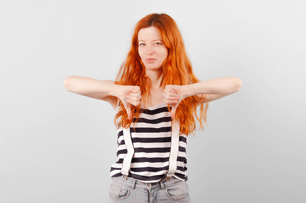 Redhead girl frowns, squints and shows thumb down. Negative and Condemnation - Fotoğraf, Görsel
