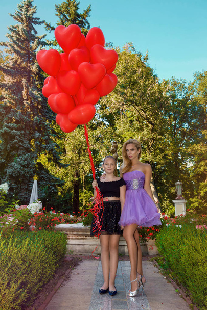 Valentine Beautiful young mother and daughter with red balloons laugh, in the park. Beautiful happy mom and kid. Birthday party. Joyful models posing, having fun, celebrating Valentine's Day - picture - Photo, Image