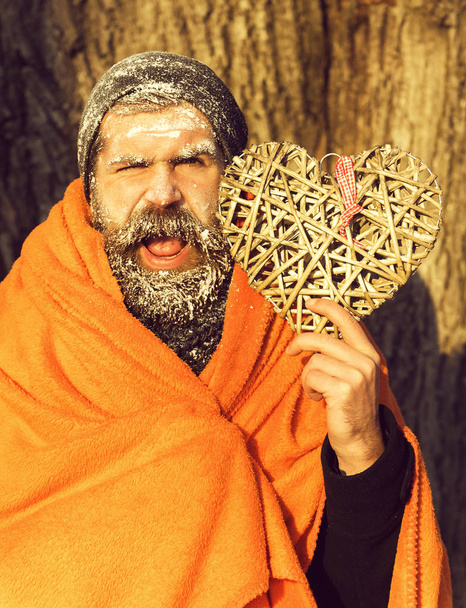 Winking frozen man, bearded hipster, with beard and moustache covered with white frost wrapped in orange blanket with wicker heart on winter day outdoors on natural backgroun - Fotoğraf, Görsel