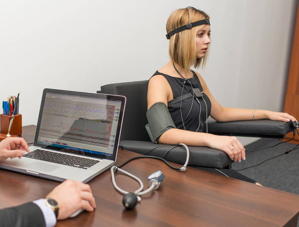 A woman with light hair dressed in a black dress on which the polygraphs on the desk are dressed are polygraph, laptop and devices - Photo, Image