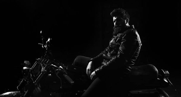 Man with beard, biker in leather jacket sitting on motor bike in darkness, black background. Macho, brutal biker in leather jacket riding motorcycle at night time, copy space. Night racer concept - Zdjęcie, obraz