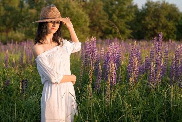 junges hübsches reizvolles Mädchen in einem purpurfarbenen Weidenhut und in einem weißen langen Kleid, das bei Sonnenuntergang auf dem Lupinenfeld steht - Foto, Bild