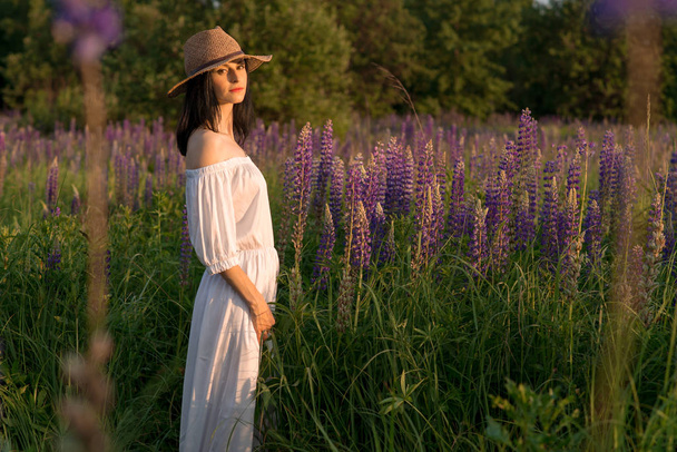 docela sexy dívka v ženském crimson proutěný klobouk a dlouhé bílé šaty stojící při západu slunce na poli Lupina - Fotografie, Obrázek