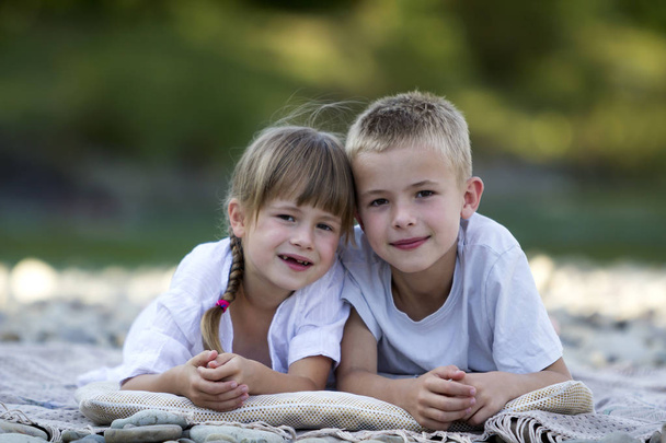 Two young happy cute blond children, boy and girl, brother and sister laying on pebbled beach on blurred bright sunny summer day background. Siblings friendship, leisure and perfect holiday concept. - Fotoğraf, Görsel