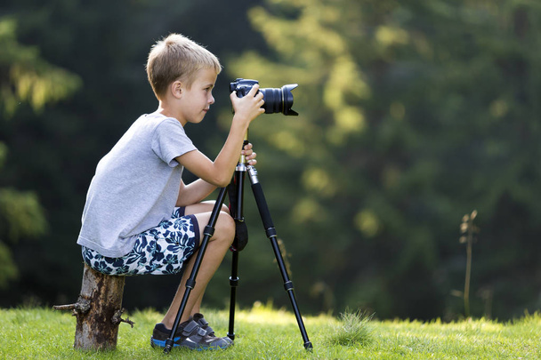 Young blond child boy sitting on tree stump on grassy clearing taking picture with tripod camera. - Photo, Image