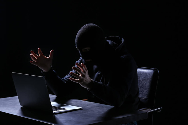 Emocjonalne haker z laptopa siedząc przy stole na ciemnym tle - Zdjęcie, obraz