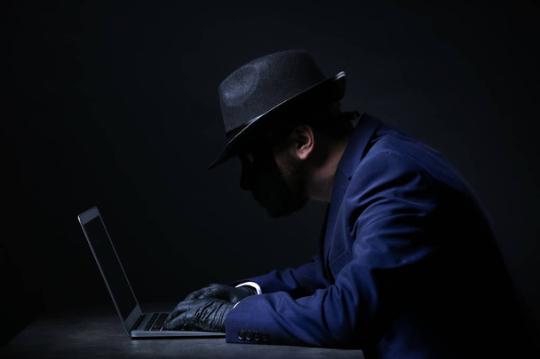 Professional hacker with laptop sitting at table on dark background - Photo, Image