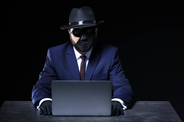 Professional hacker with laptop sitting at table on dark background - Photo, Image