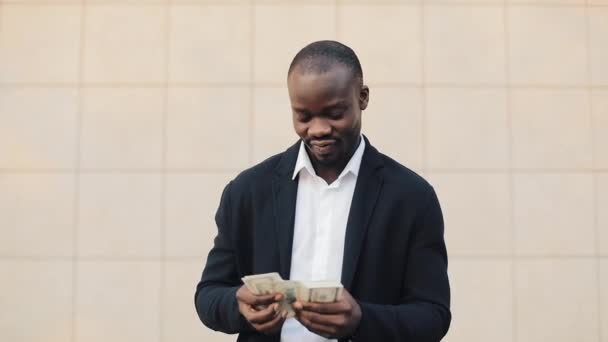 Portrait of african american businessman in suit counting money standing near office centre. He celebrating his successful win with a lot of dollars and looking into the camera - Imágenes, Vídeo