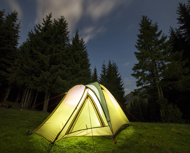 Turisztikai túrázók sátor fényesen kivilágított, a belsejében a zöld füves erdei tisztás, többek között a magas fenyőfák a távoli hegyi háttér sötét kék csillagos ég alatt. Nyáron, kemping a hegyekben éjszaka. - Fotó, kép